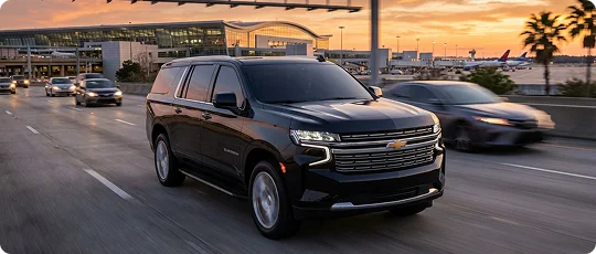 A black Chevrolet Suburban drives along a multi-lane highway toward a large, glass-walled airport terminal at dusk.