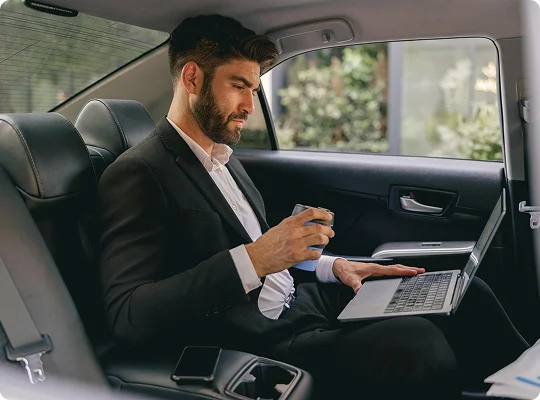 Focused executive in a black suit working on a laptop and holding a drink while seated comfortably in a black leather backseat.