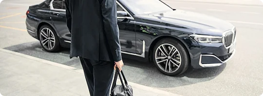 A businessman in a suit approaches a sleek BMW 7 Series sedan waiting at the curb on a bright day.