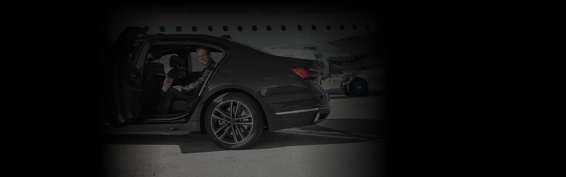 Business traveler smiling while seated in a luxury black sedan parked on the tarmac, showcasing Atlanta Airport Car Service with a clear side-angle view near a private jet.